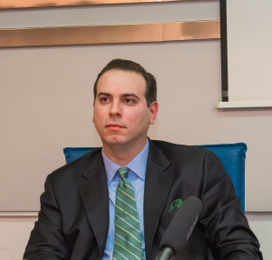 Portrait of Dr. Filip Ivanovic. He is wearing a suit, a green tie, and is sitting in front of a microphone.